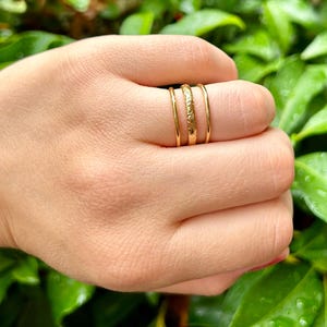 May include: Three gold rings stacked on a finger. The top ring has a textured surface, the middle ring is smooth, and the bottom ring is smooth.
