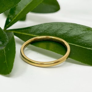 May include: A gold ring sits on a white surface, with green leaves in the background. The ring is a simple band with a polished finish. The ring is a classic design, suitable for everyday wear. The ring's diameter is approximately 2 cm.