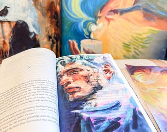 May include: An open book featuring a printed illustration of a person with blue hair and facial markings on the right page, with text on the left page. Behind the book, two colorful art prints are visible, one showing a candle and swirling blue light.