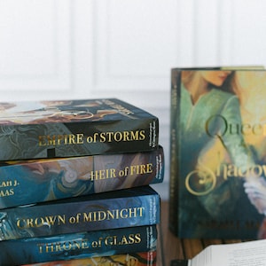 May include: A stack of fantasy novels with gold lettering on the spines. Titles include "Empire of Storms," "Heir of Fire," "Crown of Midnight," and "Throne of Glass." The covers feature colorful artwork. A book with the title "Queen of Shadows" is also visible.
