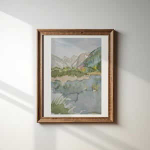 Op de afbeelding: Aquarel landschapskunst in een houten lijst. Het kunstwerk toont een serene bergscène met een meer op de voorgrond. Het kleurenpalet omvat tinten groen, blauw en bruin, wat een rustgevende esthetiek creëert. De lijst heeft een warme bruine kleur.
