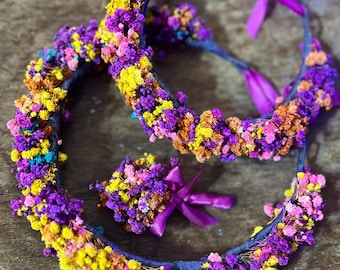 May include: Two floral headbands and a small bouquet. The headbands are adorned with colorful dried flowers in shades of purple, yellow, orange, pink, and blue. The bouquet has a matching purple ribbon.