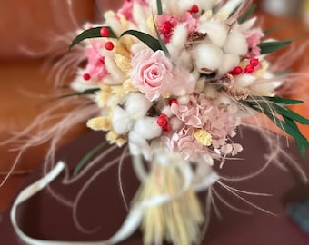 May include: A close-up of a bridal bouquet featuring a light pink rose, cotton bolls, and dried flowers. The bouquet includes pink hydrangeas, red berries, and green leaves, tied with a white ribbon. The arrangement has a soft, natural aesthetic.