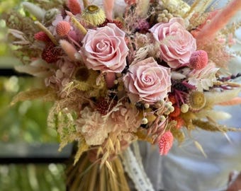May include: A bridal bouquet featuring soft pink roses and a variety of dried flowers and grasses in shades of pink, cream, and beige. The stems are wrapped with natural twine and accented with a delicate lace ribbon.