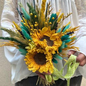 Puede incluir: Un vibrante ramo con grandes girasoles amarillos como punto focal, complementado con flores y hierbas secas de color turquesa, amarillo y verde. El ramo está atado con una cinta verde claro con lunares blancos.