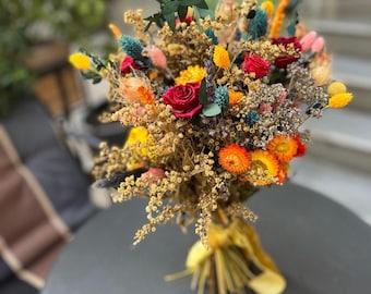 May include: A vibrant bouquet of dried flowers featuring a mix of colors. The arrangement includes red roses, yellow, orange, and blue accents, tied with a yellow ribbon. The bouquet is a mix of textures and shapes, creating a visually appealing display.
