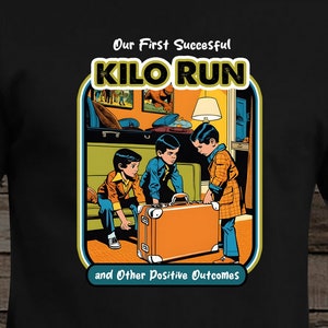 May include: Black t-shirt with a retro-style illustration of three boys attempting to lift a large suitcase. The text reads "Our First Successful KILO RUN and Other Positive Outcomes."