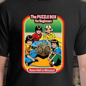 May include: A black t-shirt with a colorful graphic of a puzzle box with four children chained to it. The graphic has the text "The Puzzle Box for Beginners" and "Raise Hell in Minutes!"