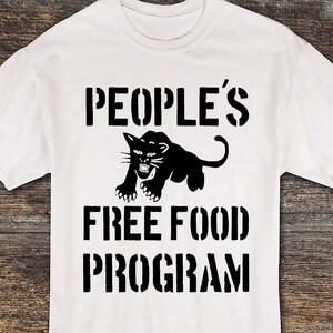 Può includere: T-shirt bianco con la scritta "PEOPLE'S FREE FOOD PROGRAM" in nero con caratteri a stencil. Una grafica di pantera nera è centrata sopra il testo. La maglietta è esposta su una superficie di legno.