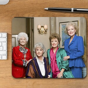 May include: A mousepad featuring a photo of four women, three of whom are wearing red, green, and blue jackets. The fourth woman is wearing a pink and green robe. The women are standing in a room with a wooden floor and white walls.