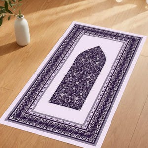 Op de afbeelding: Een rechthoekig gebedskleed met een centraal bloemmotief in donkerblauw op een lichtpaarse achtergrond. Het kleed heeft een donkerblauwe rand met ingewikkelde patronen. Het kleed ligt op een houten vloer, met een vaas en boeken op de achtergrond.