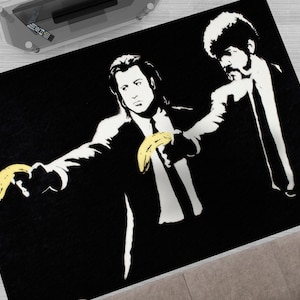 Puede incluir: Alfombra de arte de esténcil en blanco y negro con dos figuras, una sosteniendo un plátano amarillo, con el texto "Say What?" en blanco.