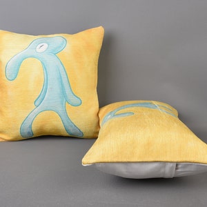 May include: Two decorative throw pillows. One pillow is yellow with a blue figure of a person with a long nose. The other pillow is yellow.