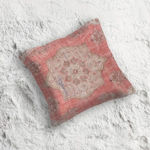 以下が含まれることがあります： 色あせたヴィンテージスタイルのデザインの正方形の装飾枕。枕は、伝統的なラグを連想させる、コーラル、ベージュ、くすんだブルーの色合いの、中央のメダリオンパターンが特徴です。枕はふわふわの白い表面に置かれています。