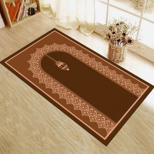 May include: A brown prayer rug with a beige border and a decorative arch design featuring a hanging lantern. The rug has a soft, plush texture and is perfect for use in prayer.