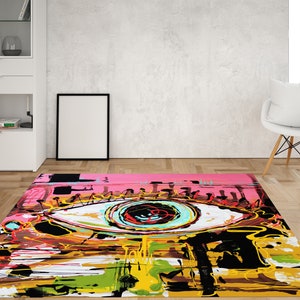 May include: Abstract rug with a large, colorful eye design. The eye is painted in shades of blue, green, yellow, red, and black. The background is a pink and yellow abstract design.