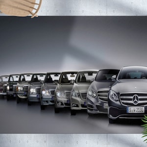 Op de afbeelding: Een rij Mercedes-Benz E-Klasse sedans, die de evolutie van het model laat zien van de jaren 1970 tot heden. De auto's zijn in chronologische volgorde gerangschikt, met de oudste auto links en de nieuwste auto rechts. De nieuwste auto heeft een kentekenplaat met de tekst "S.W 2130".