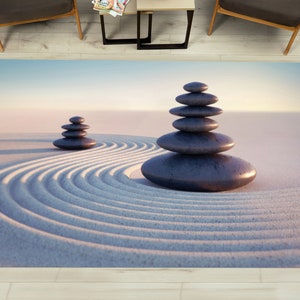 Puede incluir: Una alfombra de jardín zen con fondo de arena blanca y dos pilas de piedras grises. La arena tiene un patrón circular.
