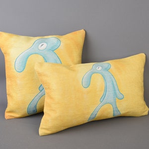 May include: Two yellow throw pillows with a blue cartoon figure printed on each. The figure is a stick figure with a long neck and a single eye.