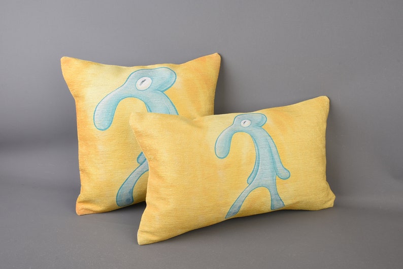 May include: Two yellow throw pillows with a blue cartoon figure printed on each. The figure is a stick figure with a long neck and a single eye.