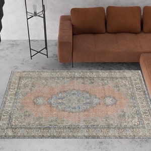 Könnte beinhalten: Ein Vintage-Teppich mit einem verblassten rosa und blauen Blumenmuster. Der Teppich hat einen hellbraunen Rand mit komplizierten Details. Der Teppich befindet sich in einem Wohnzimmer mit einem braunen Ledersofa.