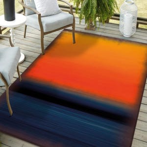 Pode incluir: Um tapete retangular para exterior com um design abstrato. O tapete apresenta um gradiente de cores, que transita do laranja para o vermelho e depois para o azul escuro. O tapete está colocado num deck de madeira, sugerindo um espaço de convívio ao ar livre.
