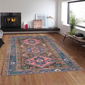 Könnte beinhalten: Ein rechteckiger Teppich mit einem aufwendigen geometrischen Muster. Der Teppich hat ein zentrales Design in Braun-, Rosa- und Blautönen, umgeben von einem blauen und beigen Rand. Er liegt auf einem Holzboden.