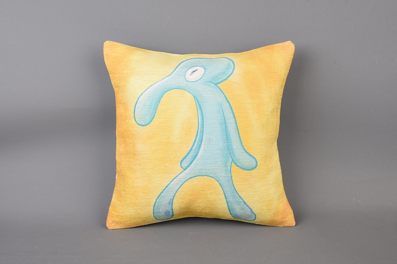 May include: A yellow throw pillow with a blue, cartoon-like figure of a person with a long nose and arms. The figure is standing on the pillow.