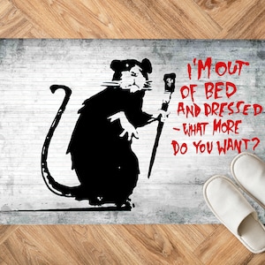 Puede incluir: Una ilustración en blanco y negro de una rata sosteniendo un palo con una estrella en él. La rata está de pie sobre una pared de ladrillo blanco con el texto "I'm out of bed and dressed - what more do you want?" en rojo.