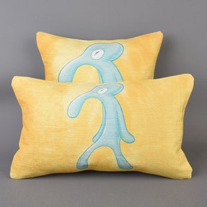 May include: Two yellow throw pillows with a blue cartoon figure printed on them. The figure is a stick figure with a long nose and a single eye.