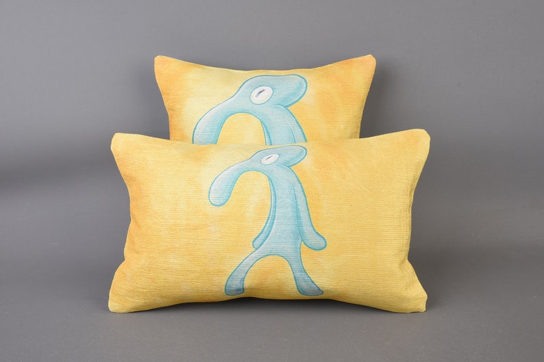 May include: Two yellow throw pillows with a blue cartoon figure printed on them. The figure is a stick figure with a long nose and a single eye.