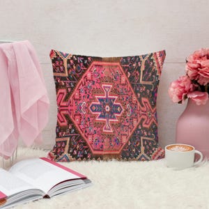 May include: A decorative throw pillow with a geometric pattern in shades of pink, brown, and blue. The pillow has a textured appearance and is placed on a white, fluffy surface. A pink vase with flowers and a cup of coffee are nearby.