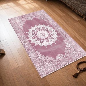 May include: A rectangular prayer rug with a white floral design on a mauve background. The rug is placed on a wooden floor, with a wooden book stand and prayer beads nearby. The rug has a decorative border.