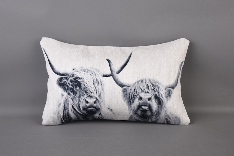 May include: A white rectangular cushion with a black and white image of two Highland cattle. The cattle are looking directly at the viewer. The cushion is made of a soft, textured fabric.