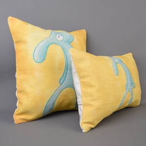 May include: Two yellow throw pillows with a blue cartoon character printed on them. The character is a stick figure with a long neck and a single eye. The pillows are made of a soft, textured fabric.