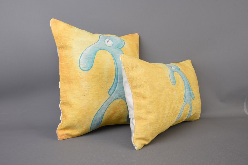 May include: Two yellow throw pillows with a blue cartoon character printed on them. The character is a stick figure with a long neck and a single eye. The pillows are made of a soft, textured fabric.