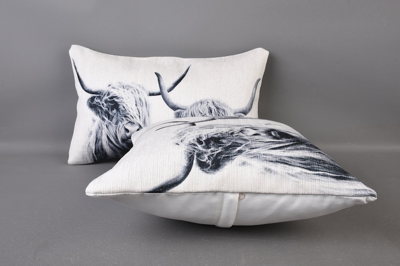 May include: Two white throw pillows featuring a black and white image of a Highland cow on each. The pillows are made from a soft, textured fabric.