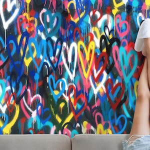 May include: A colourful wall mural featuring a pattern of hearts in various colours, including pink, blue, yellow, red, and white. The hearts are painted in a graffiti style with drips and splatters. A person's legs are visible at the bottom of the image, wearing white slippers and denim shorts.