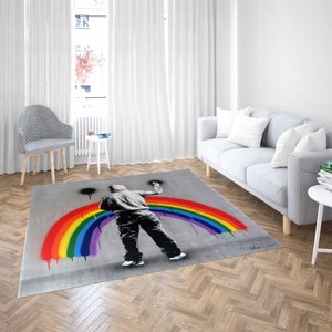Könnte beinhalten: Ein grauer Teppich mit einem darauf gemalten Regenbogen. Eine schwarze Figur malt den Regenbogen mit einer Sprühdose.