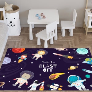 Puede incluir: Una alfombra colorida con temática espacial con fondo azul oscuro, estrellas, planetas y cohetes. La alfombra presenta un león, un oso y un astronauta de dibujos animados. El texto "3...2...1... BLAST OFF" está impreso en la alfombra.