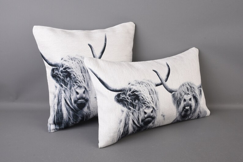 May include: Two white cushion covers with black and white images of Highland cattle. The cushions are on a grey background.