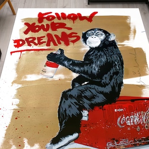 May include: A black and white illustration of a chimpanzee holding a spray can, sitting on a red cooler with the Coca-Cola logo. The background is a gold and white abstract design with the words "Follow Your Dreams" in red.