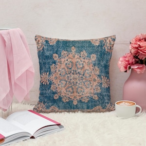 May include: Square decorative pillow with a distressed, vintage-style design. The pillow features a teal background with a central medallion pattern in shades of peach and beige. The pillow is placed on a white, fluffy surface.