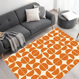 May include: A white and orange geometric area rug with a repeating pattern of semi-circles. The rug is on a hardwood floor in front of a gray sofa.