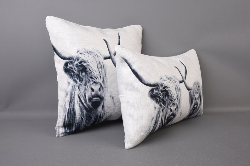 May include: Two decorative throw pillows featuring a black and white image of a Highland cow's face. The pillows are made from a white fabric with a subtle texture.