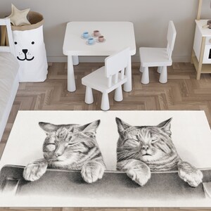 May include: A white area rug with a black and white illustration of two cats sleeping. The cats are drawn in a realistic style with their eyes closed and their paws resting on a surface.