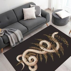 Op de afbeelding: Een zwart en gouden tapijt met een wit slangenontwerp. De slang is om een ​​varenblad gewikkeld. Het tapijt is perfect om een ​​touch van natuur aan uw huis toe te voegen.