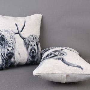 May include: Two white and black throw pillows featuring a printed image of Highland cattle. The pillows are made from a soft, textured fabric.