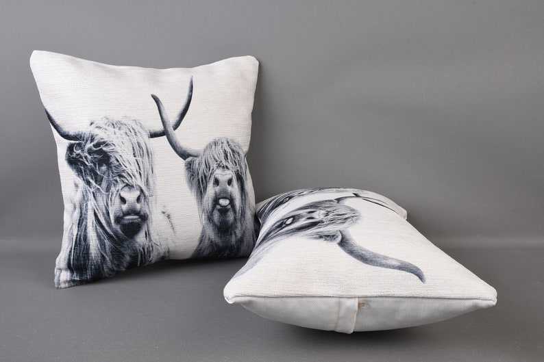 May include: Two white and black throw pillows featuring a printed image of Highland cattle. The pillows are made from a soft, textured fabric.