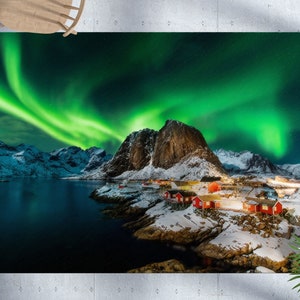May include: A scenic landscape photograph featuring a vibrant green aurora borealis over a snow-covered mountain range and a small village with red houses. The water in the foreground is a deep blue, reflecting the light of the aurora.